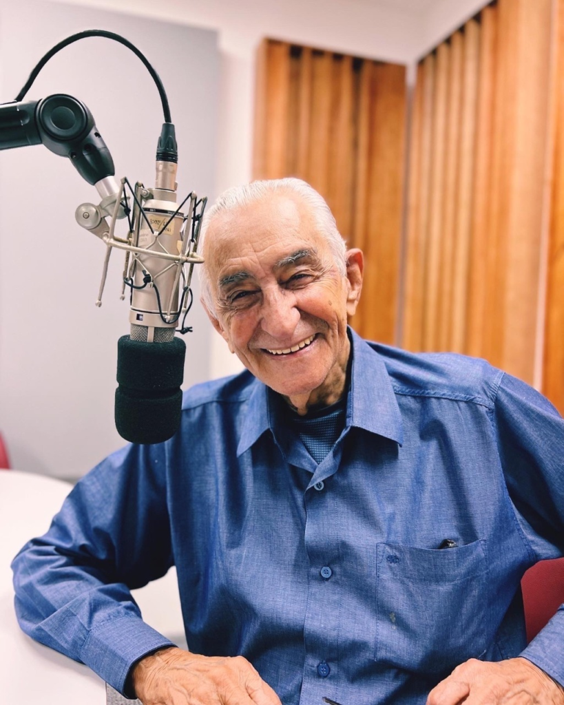 Ricardo Parreiras, cabelos grisalhos, sorridente e usando camisa social azul, sentado em frente a um microfone na Rádio Inconfidência. Ao fundo, paredes revestidas com ripas de madeira.