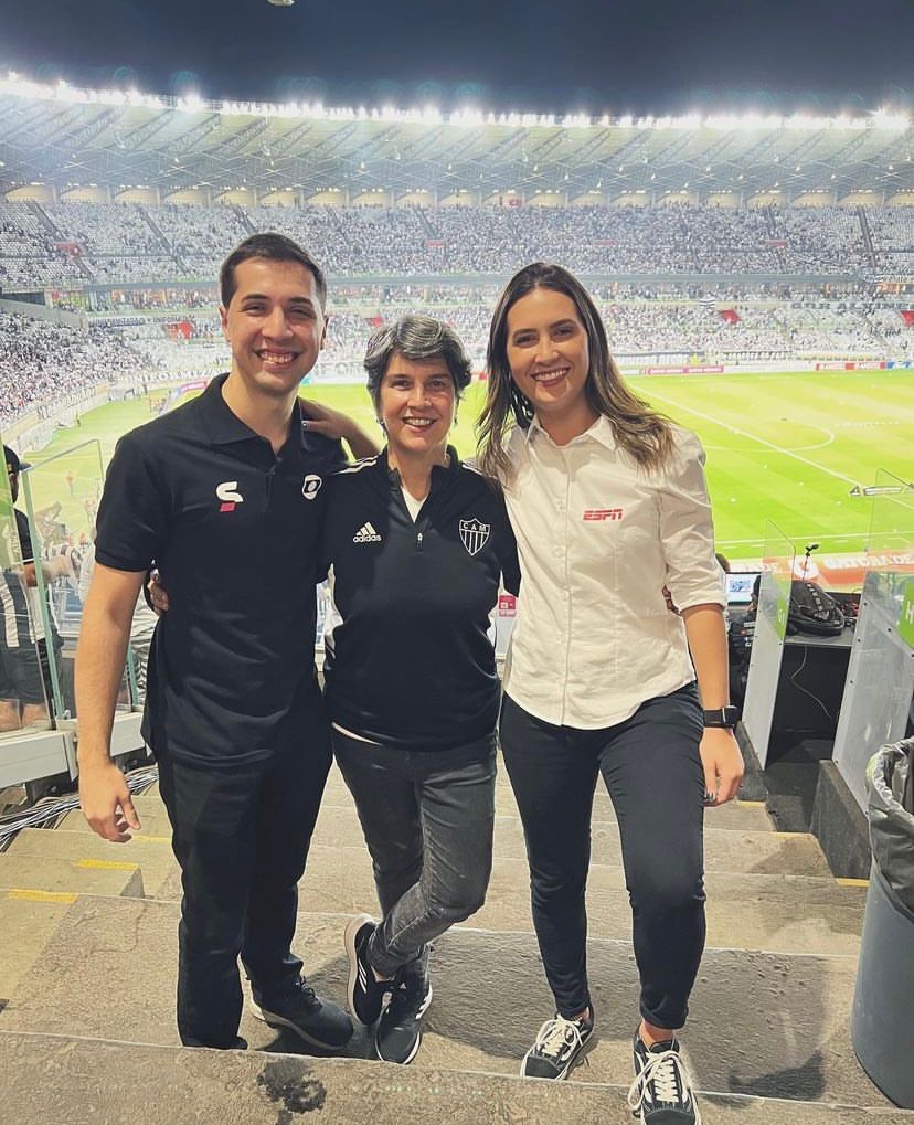 Pedro (à esquerda) com a mãe Adriana (ao centro) e a irmã Mariana (à direita), abraçados e em pé em escada do Estádio Mineirão, em cobertura de partida de futebol, com o campo e a torcida ao fundo. Adriana usa camisa do Atlético Mineiro e Pedro e Mariana usam, respectivamente, os uniformes da SporTV e da ESPN.
