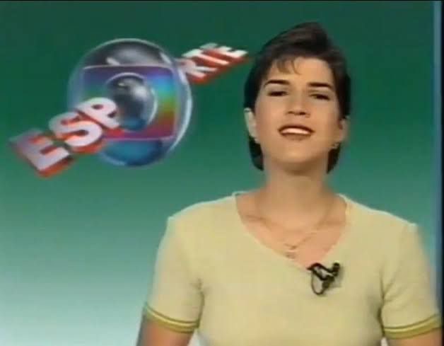 Adriana Spinelli apresentando o Globo Esporte, em primeiro plano, com cabelo castanho e curto, blusa bege, microfone de lapela em fundo verde com logo do Globo Esporte no canto superior esquerdo.
