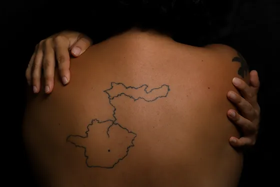 
Foto com fundo preto mostrando apenas as costas de Joana, com uma tatuagem dos estados de Pernambuco e Minas Gerais unidos pela linha que representa o rio São Francisco. Ela está com as mãos abraçando os ombros.
