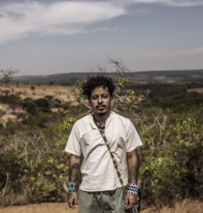 Emanuel Kaauara se encontra no centro da imagem com uma paisagem do cerrado brasileiro ao fundo. A vegetação é baixa, a terra é árida e o céu está nublado.