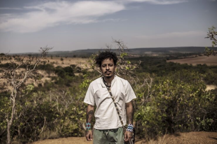 Emanuel Kaauara se encontra no centro da imagem com uma paisagem do cerrado brasileiro ao fundo. A vegetação é baixa, a terra é árida e o céu está nublado.