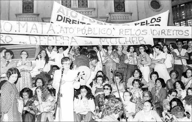 Imagem em preto e branco de manifestação contra o feminicídio. Ao fundo, a Igreja São José. Mulheres, em sua maioria brancas, levantam faixas de protesto.