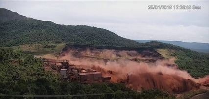 Imagem aérea do rompimento de uma barragem localizada em Brumadinho, onde é possível denotar a queda de paredes de lama e barro destruindo sítios de construção.