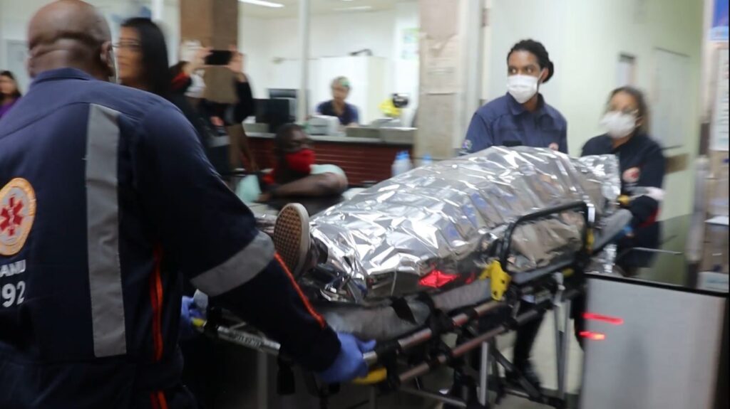 Paciente envolto em proteção é levado em uma maca por dois socorristas do SAMU para o interior do Hospital João XXIII