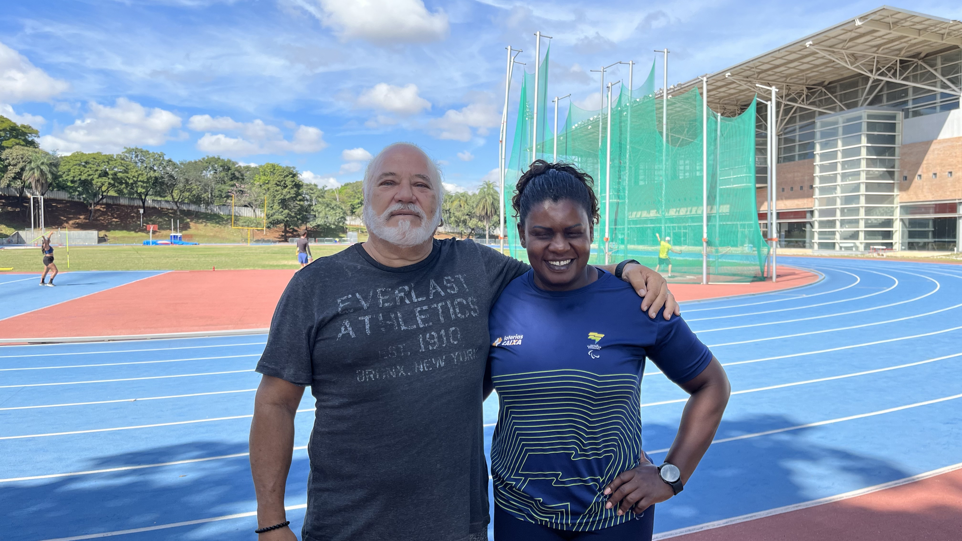 A imagem é composta por um céu azul e o centro de treinamento da Universidade Federal de Minas Gerais. No meio da imagem, um homem branco de cabelos grisalhos abraçada com uma mulher negra que é atleta paralímpica. 