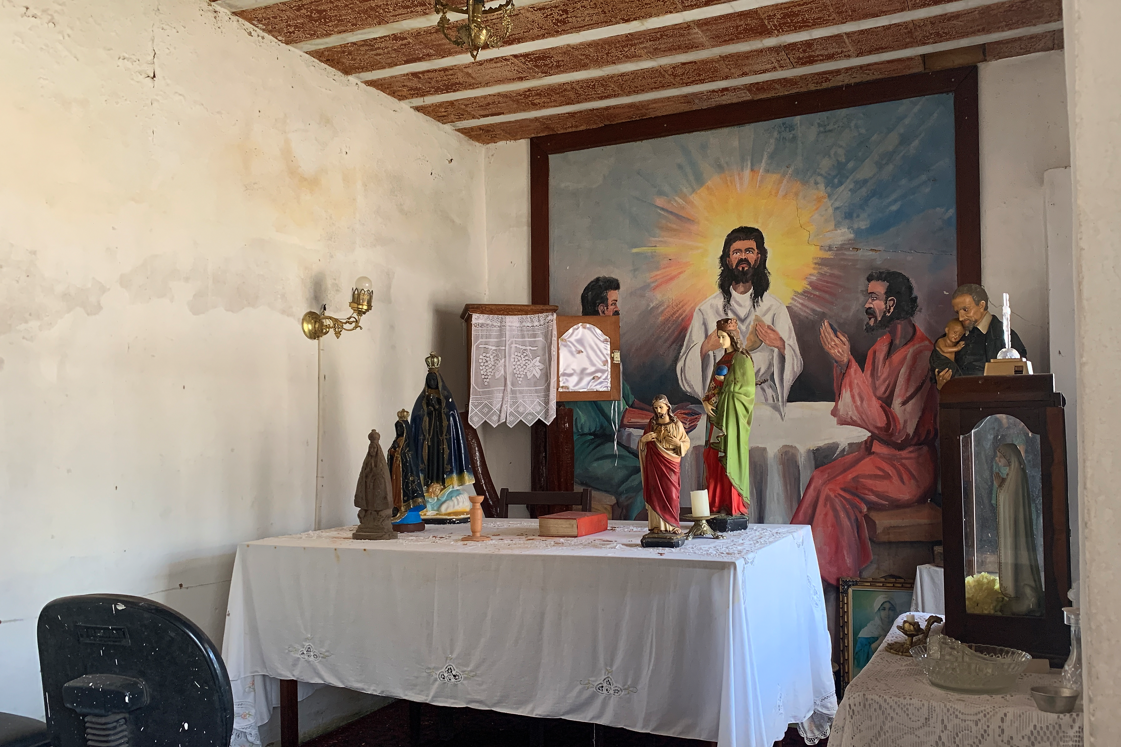 Fotografia digital de uma sala com figuras religiosas e uma imagem de Jesus Cristo ao fundo. Há infiltrações nas paredes e tijolos aparentes no teto.