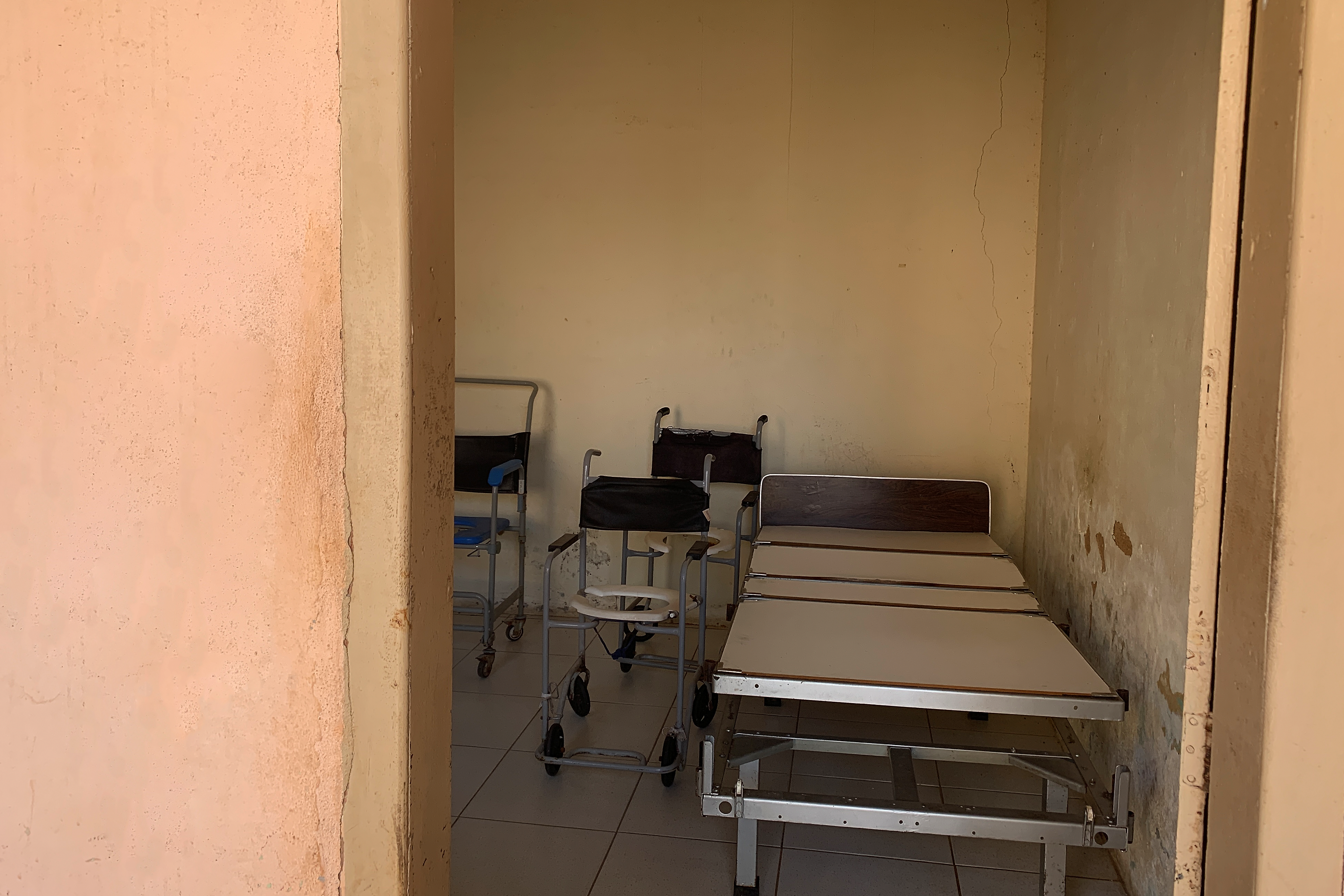 Fotografia digital colorida. Atrás de uma porta, vemos materiais inutilizados, como cadeiras de rodas e cama hospitalar.