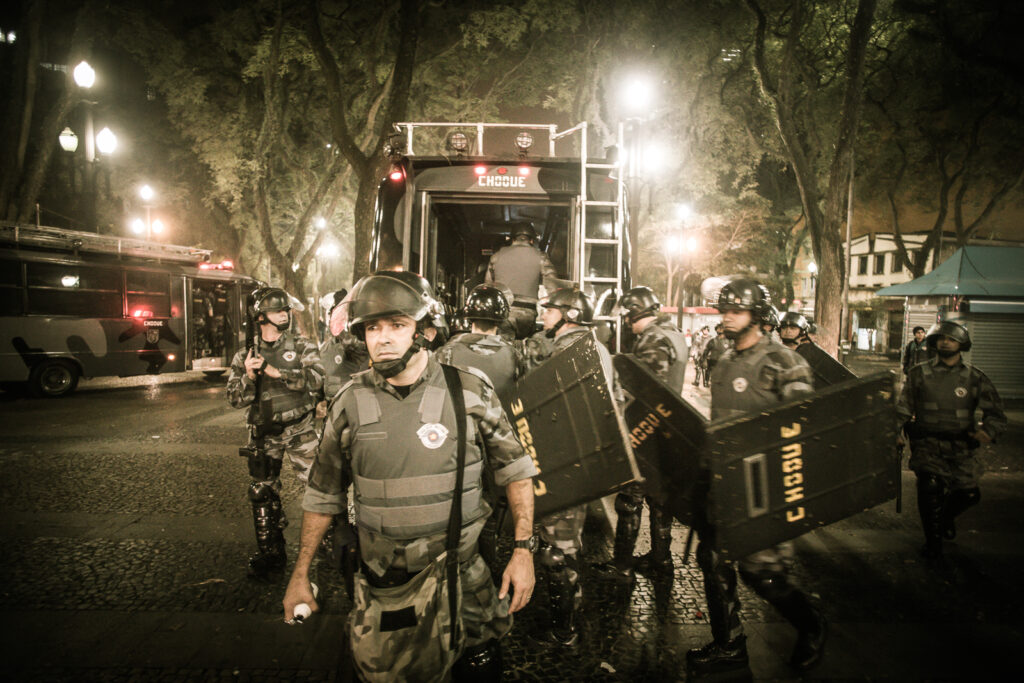 Foto colorida tirada durante a noite, estão em um ambiente aberto, com árvores e prédios ao fundo, veículos com a camuflagem cinza e um grupo de Policiais Militares da Tropa de Choque de São Paulo em um momento de concentração para ações nas manifestações de junho de 2013.