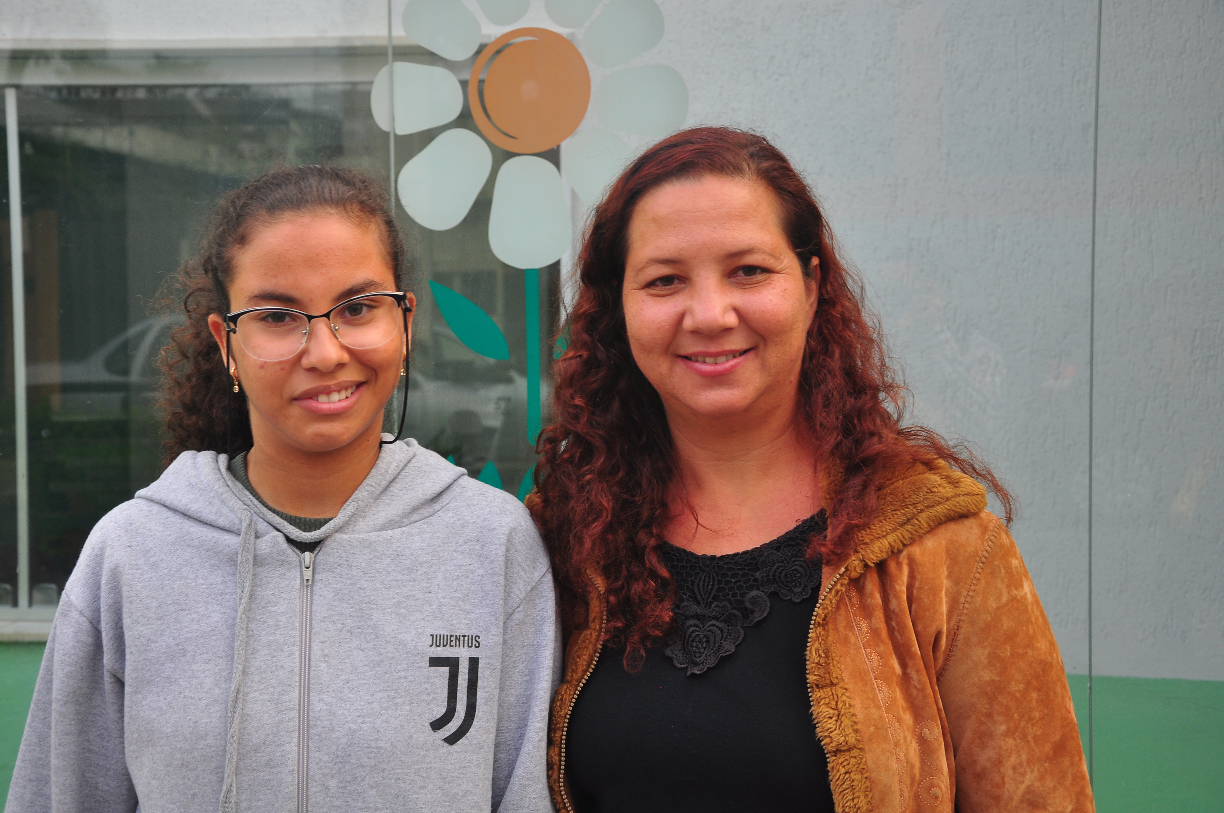 Maria Vitória e a mãe, Gilmara Junia Campos posadas uma ao lado da outra par uma foto em frente à fachada da APAE, clínica onde Maria Vitória tem acompanhamento médico e social. As duas sorriem.