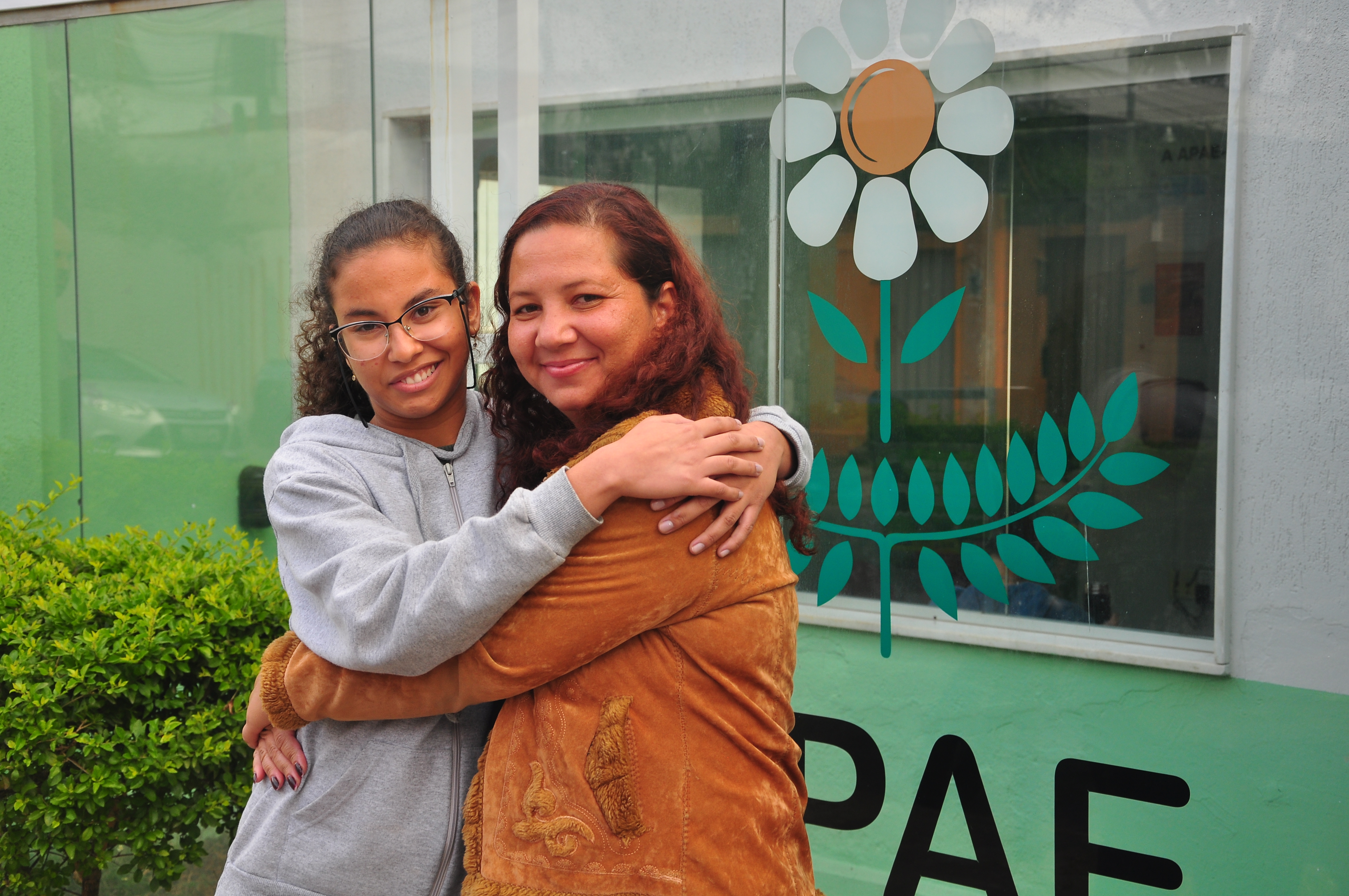 Maria Vitória e a mãe, Gilmara Junia Campos posadas uma ao lado da outra para uma foto em frente à fachada da APAE, clínica onde Maria Vitória tem acompanhamento médico e social. Legenda abaixo: Maria Vitória é acompanhada na APAE BH há 2 anos.