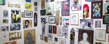 Fotografia colorida de uma galeria com paredes brancas cobertas por imagens, pinturas e peças artísticas que remetem a pichação, é possível ver também um chão de tacos de madeira.
