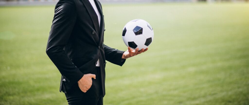 Uma figura masculina de terno preto segurando uma bola de futebol em uma de suas mãos, ele está em um fundo verde desfocado que remete a um campo de futebol.