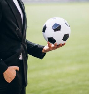 Uma figura masculina de terno preto segurando uma bola de futebol em uma de suas mãos, ele está em um fundo verde desfocado que remete a um campo de futebol.