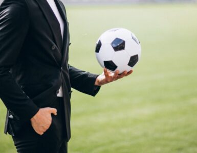 Uma figura masculina de terno preto segurando uma bola de futebol em uma de suas mãos, ele está em um fundo verde desfocado que remete a um campo de futebol.
