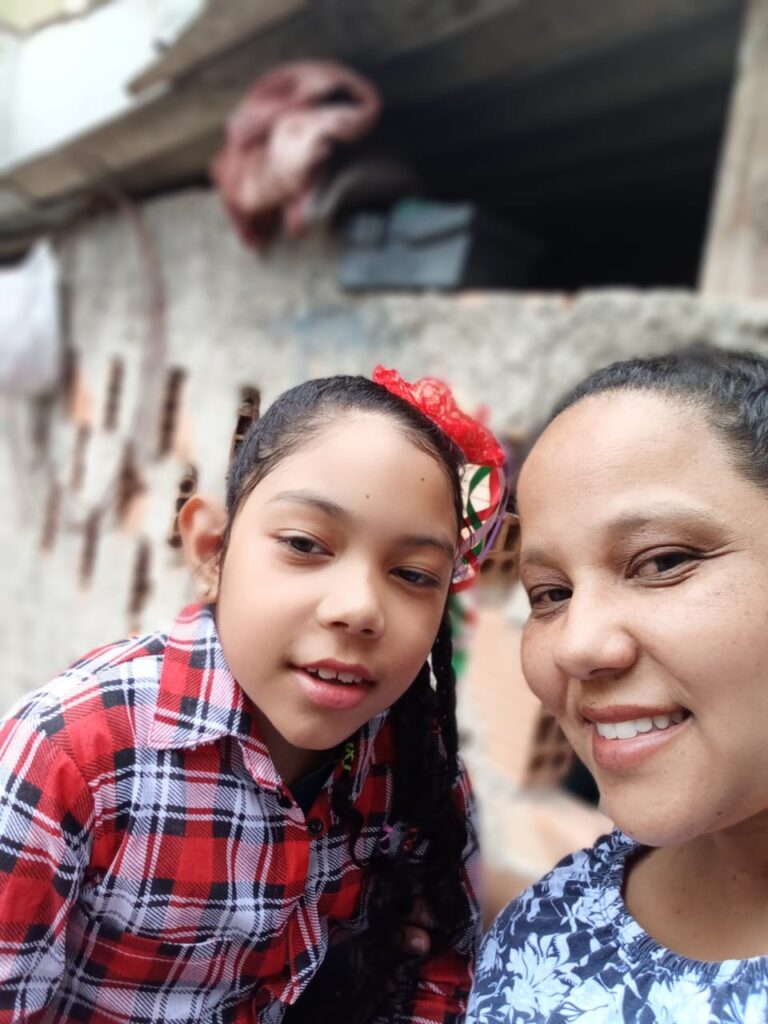 Cristiane, uma mulher parda sorri ao lado da filha, Julia. A menina está vestida com uma blusa de estampa xadrez vermelha, branco e preta, e um laço vermelho no cabelo. 