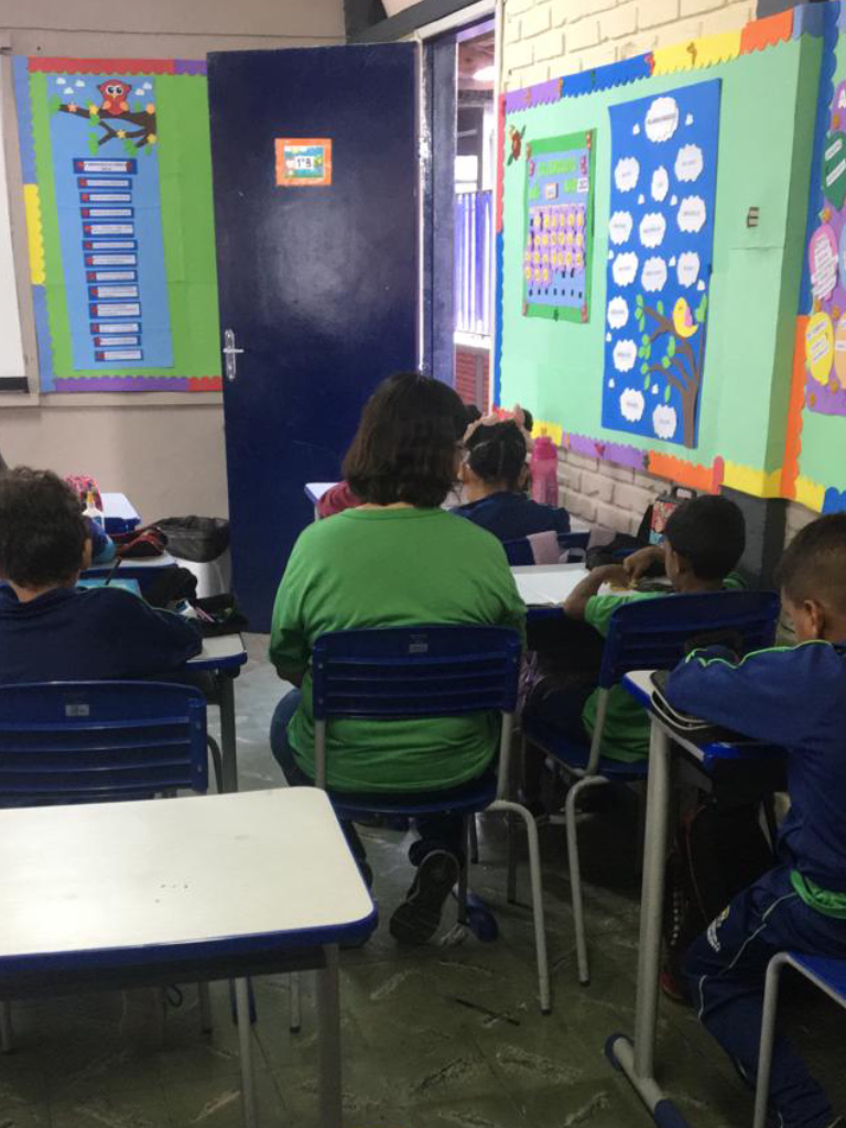 Mulher parda de cabelos escuros está sentada ao lado de um menino durante a aula. Ambos estão de costas. 
