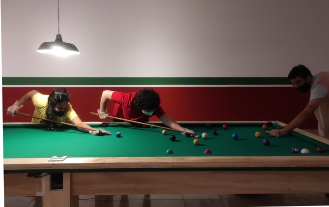 Fotografia em que três pessoas jogam sinuca em uma mesa de tamanho aumentado e com mais bolas do que o comum, ao fundo há uma parede com uma parte vermelha, uma faixa verde e outra grande parte branca, remetendo um boteco.