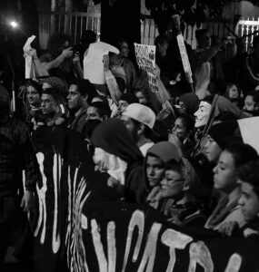 Imagem em preto e branco mostra um grupo de Black Blocs segurando uma faixa durante Jornadas de Junho de 2013