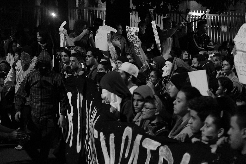 Foto em preto e branco registrando um grupo de manifestantes, denominados pelos veículos de imprensa, black blocs. O grupo é composto por homens e mulheres adultos, alguns utilizam capuz e máscaras e outros estão com o rosto descoberto. Estão em um espaço aberto, durante a noite e com um prédio de fundo, alguns estão segurando cartazes de protestos.