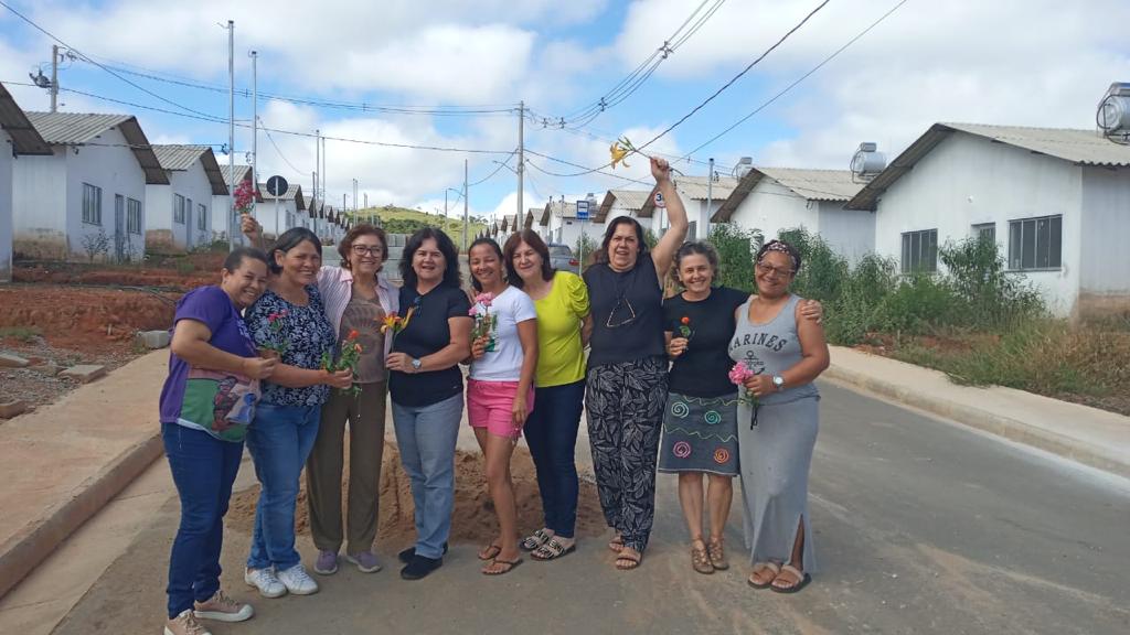 Nove mulheres, uma ao lado da outra segurando flores em suas mãos, ao fundo: casas recém construídas semelhantes umas às outras. Rua asfaltada e alguma vegetação em torno das casas.
