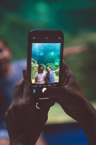 Pessoa segura um celular preto com as duas mãos enquanto tira foto de duas crianças ao fundo.
