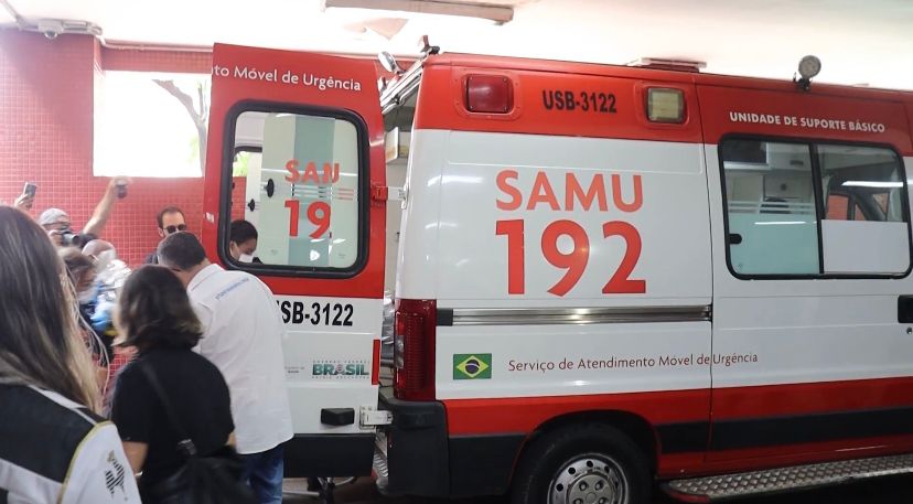 Fotografia digital colorida de uma ambulância do SAMU em frente ao hospital João XXIII. À esquerda, pessoas se aglomeram.
