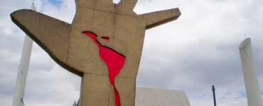 Foto colorida da escultura "Mão" de Oscar Niemeyer. Em primeiro plano, a escultura que mostra uma mão para cima com os dedos abertos e com o mapa da América Latina escorrendo sangue na palma da mão