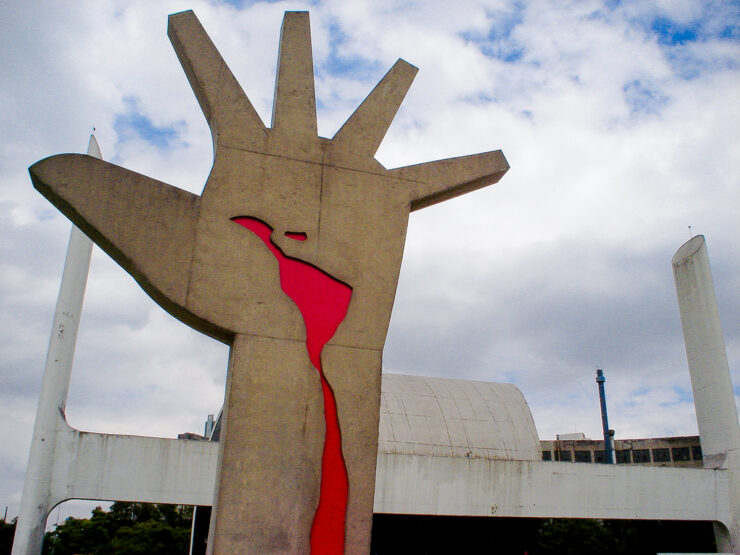 Foto colorida da escultura "Mão" de Oscar Niemeyer. Em primeiro plano, a escultura que mostra uma mão para cima com os dedos abertos e com o mapa da América Latina escorrendo sangue na palma da mão