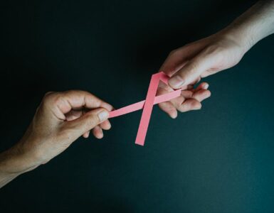 Imagem de laço rosa, símbolo da campanha outubro rosa, sendo repassado de uma mão para outra.