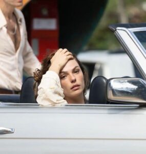 A personagem Ângela Diniz, uma mulher branca, com o cabelo castanho, está dentro de um carro conversível prata, no banco do motorista. Sua mão esquerda sobre a cabeça e seu olhar é distante.