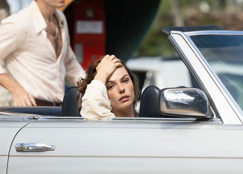 A personagem Ângela Diniz, uma mulher branca, com o cabelo castanho, está dentro de um carro conversível prata, no banco do motorista. Sua mão esquerda sobre a cabeça e seu olhar é distante.