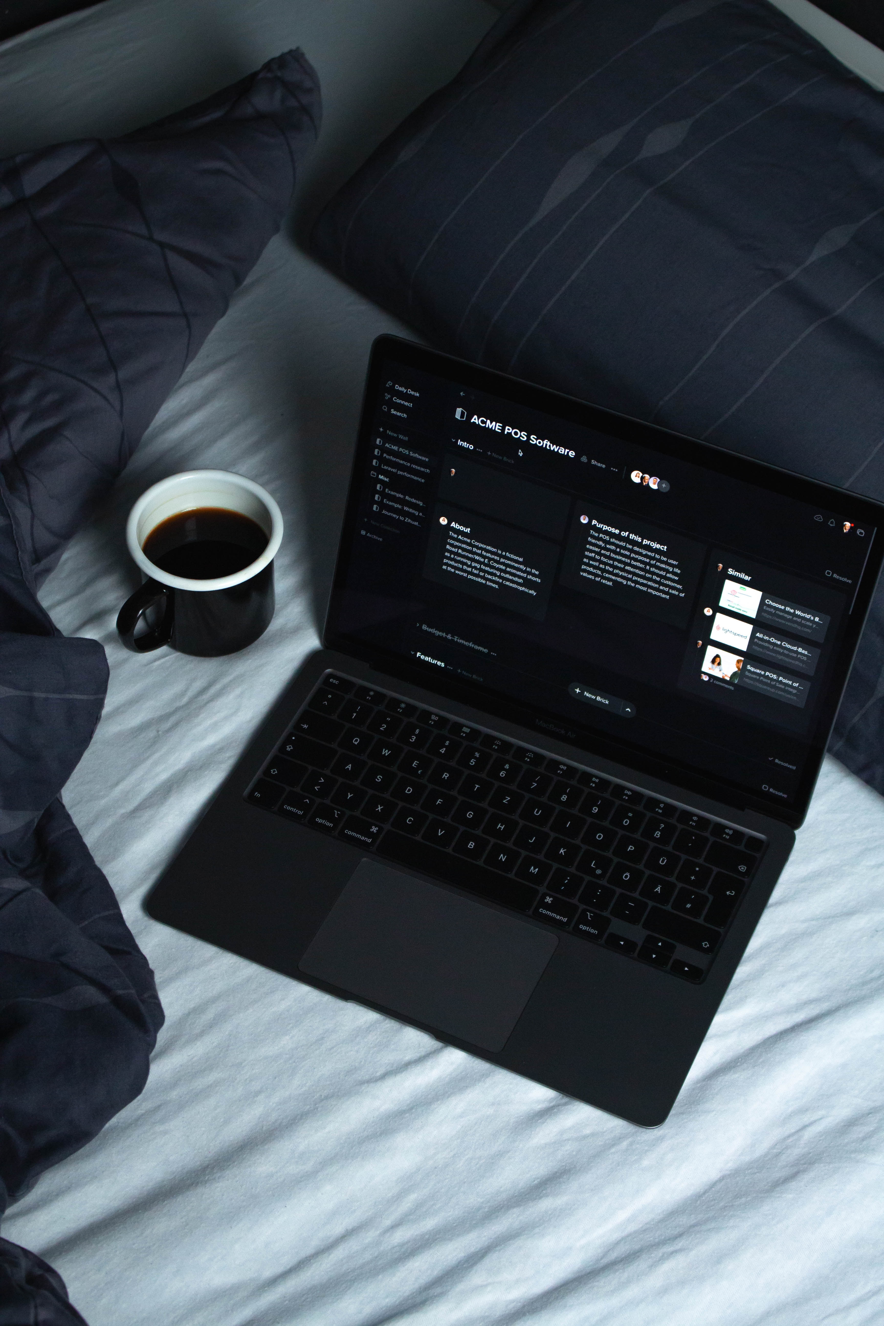 Notebook com tela de agenda de trabalho aberta, posicionado em cima da cama ao lado de uma xícara preta contendo café dentro
