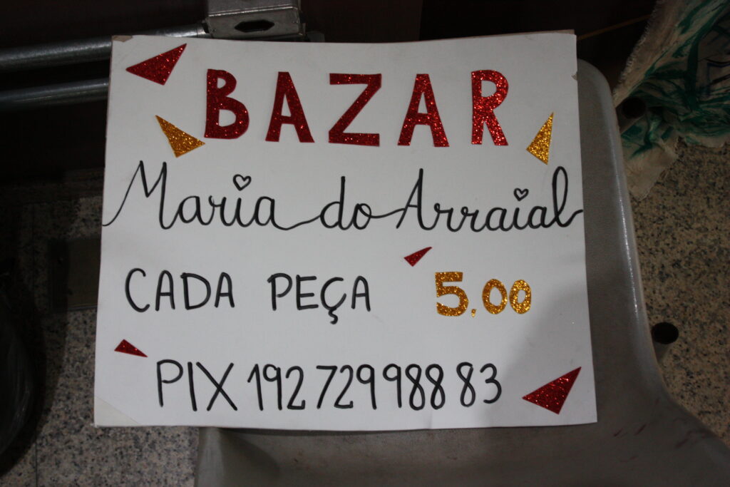 O cartaz é feito com uma folha branca e folhas de EVA com purpurina nas cores amarelo e vermelho foram cortadas para fazer triângulos que ficam espalhados pelo cartaz, a palavra "Bazar" e o número "5,00". No cartaz lê-se: "Bazar Maria do Arraial/ Cada peça 5,00/ Pix 19272998883".