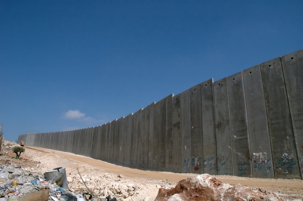 A imagem mostra um muro de concreto alto e grosso que se estende por uma paisagem árida. O muro divide área da Cisjordânia e Israel. A imagem mostra o céu azul