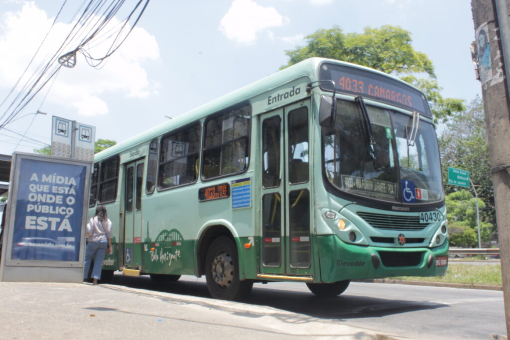 Ônibus de cor verde parado em um ponto de ônibus e prestes a arrancar, no ponto é possivél ler os dizeres: "A mídia está onde o publico está", e ao fundo da foto é possivel notar um céu limpo com algumas nuvens brancas e a copa de uma árvore esticando-se atrás do ônibus.