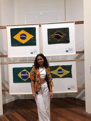 menina parda usando calça branca e um cropped branco, um kimono amarelo, possui cabelo pretos cacheados e volumosos na altura do ombro. possa de costas para uma exposição com quatro bandeiras bordadas do Brasil