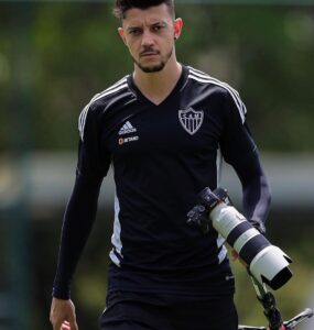 Fotógrafo Pedro Souza com uma camisa do Atlético, preta, de manga longa. Está segurando o tripé e uma câmera na mão.