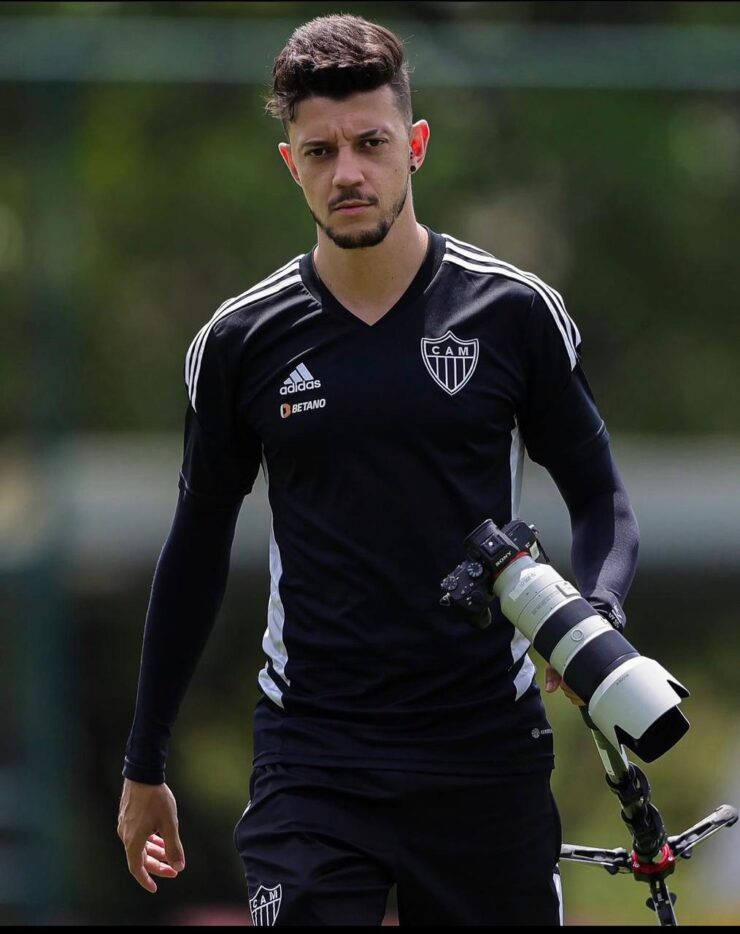 Fotógrafo Pedro Souza com uma camisa do Atlético, preta, de manga longa. Está segurando o tripé e uma câmera na mão.