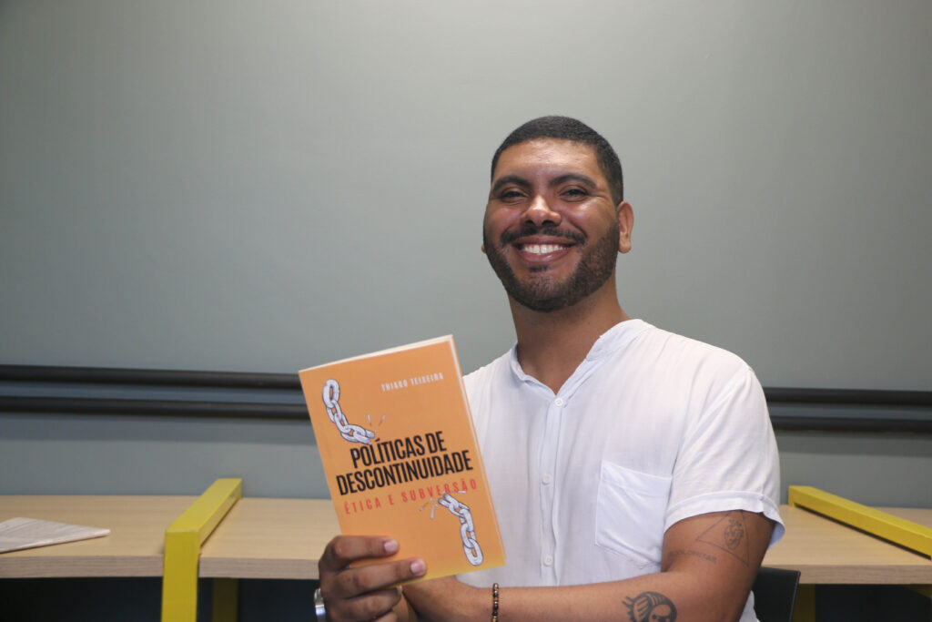 o autor Thiago Teixeira posicionado levemente à direita do centro segurando seu livro de capa alaranjada, que está ao centro próximo à margem inferior da foto. O fundo da foto é um verde acinzentado com luminosidade no meio, formando um degradê de luz. Há duas vigas cilindricas pretas coladas horizontalmente na parede, próximas a uma mesa de madeira clara dividida em sessões por duas vigar metálicas amarelas retangulares que estão uma em cada lado do Thiago.