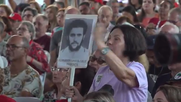 Em meio a multidão, uma mulher segura uma placa com uma fotografia em preto e branco de uma vítima da ditadura.