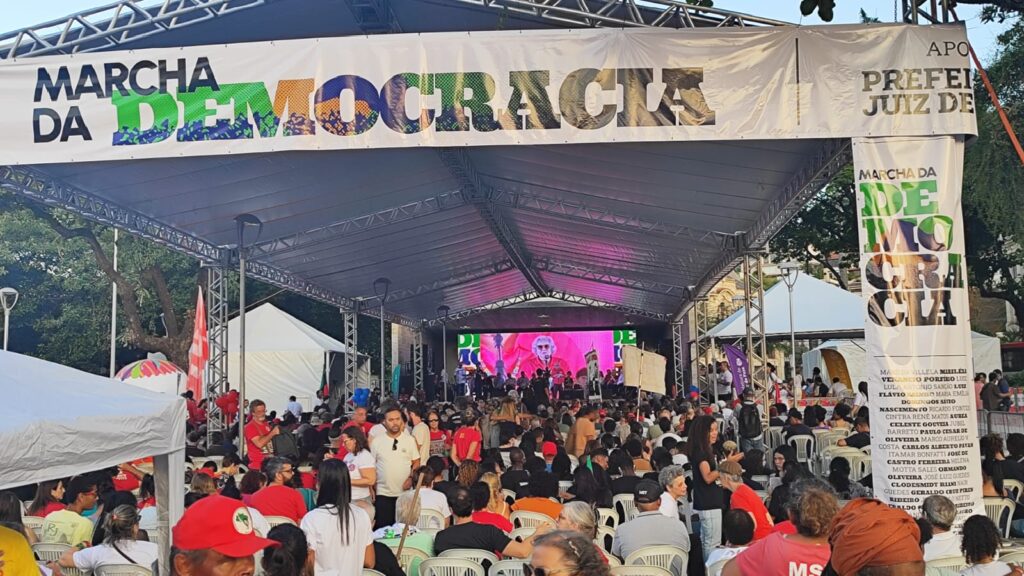 Centenas de pessoas reunidas sob uma grande tenda para acompanhar o ato em memória aos 60 anos golpe. Acima da estrutura montada há uma faixa escrito com as cores da bandeira do Brasil Marcha da Democracia