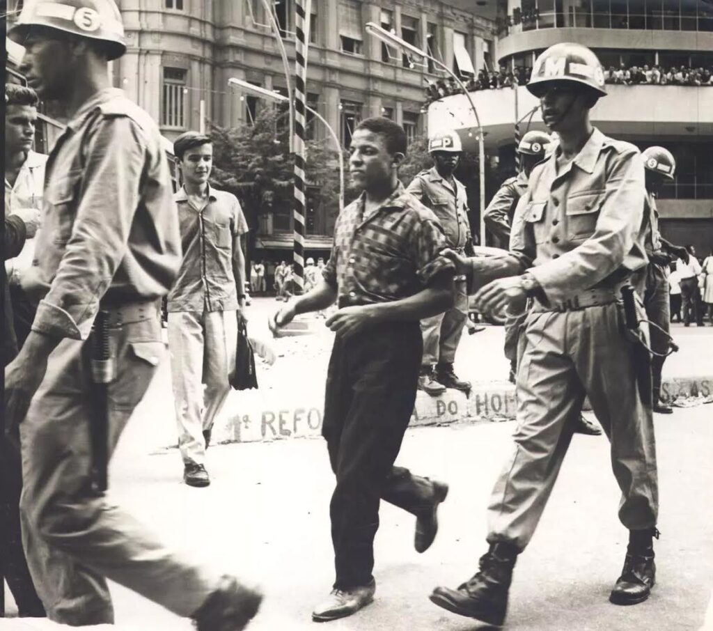Fotografia em preto e branco militares conduzindo um jovem que usa camisa estampada, calças cumpridas e sapatos fechados. 