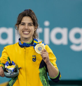 Mulher jovem branca, vestindo casaco da seleção brasileira segura medalha de ouro na mão