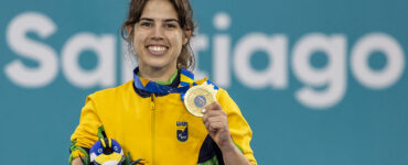 Mulher jovem branca, vestindo casaco da seleção brasileira segura medalha de ouro na mão