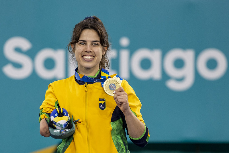 Mulher jovem branca, vestindo casaco da seleção brasileira segura medalha de ouro na mão