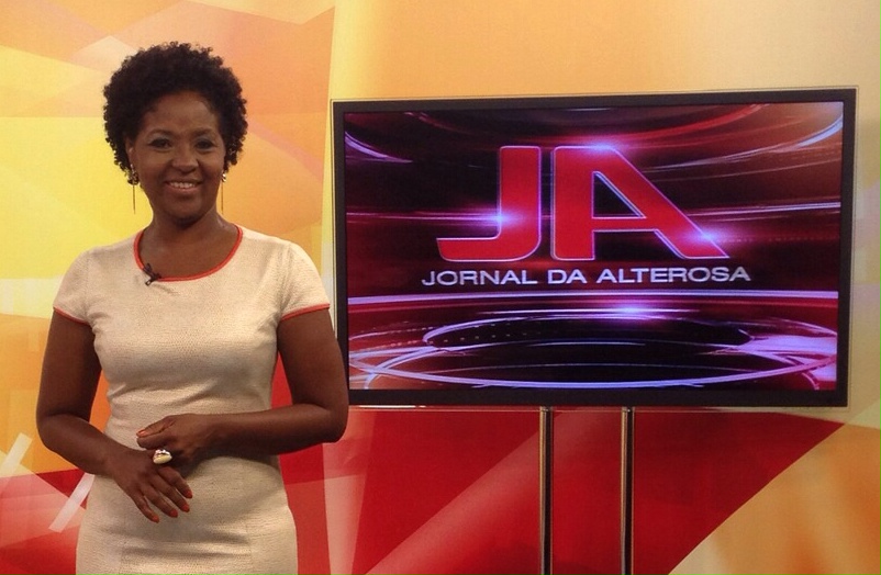 Ethel na TV Alterosa, apresentando o jornal em frente a um telão com a logo do Jornal Alterosa, com uma vestimenta branca. 