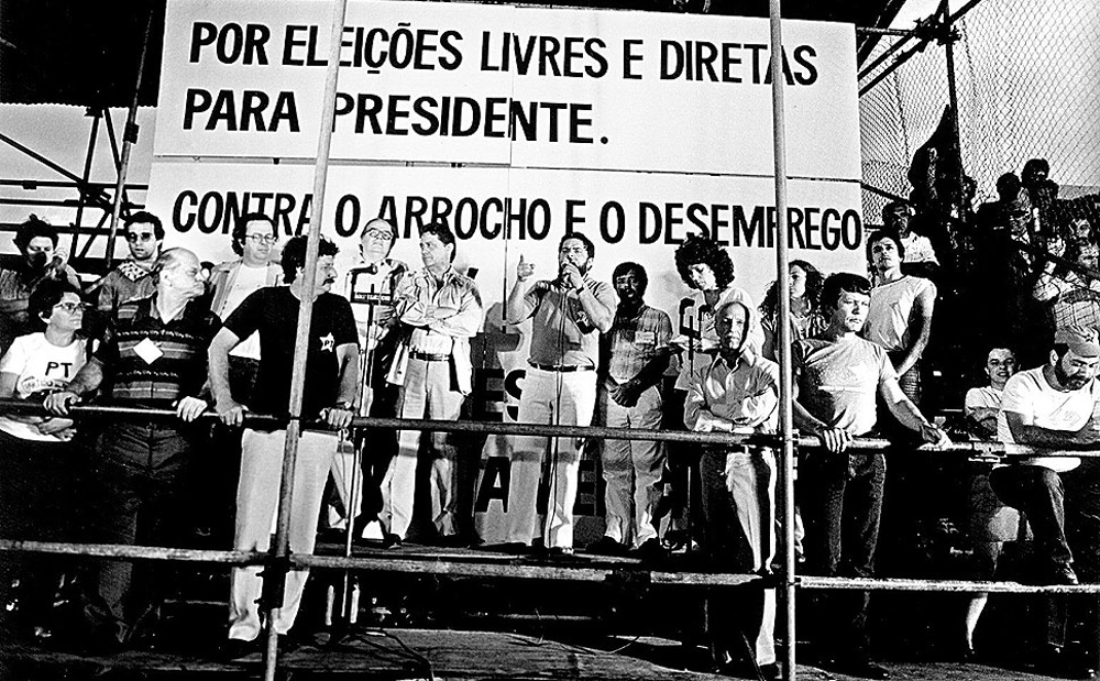 Pessoas, incluindo Lula, em um palanque. Lula está segurando o microfone. Atrás está uma placa, escrito: por eleições livres e diretas para presidente. Contra  o arrocho e o desemprego.