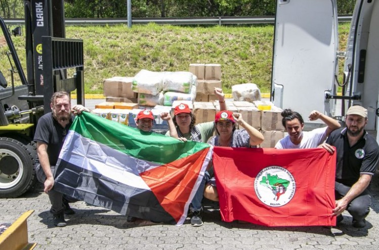 seis pessoas reunidas em frente a um caminhão com suprimentos. Elas carregam bandeiras do MST e da Palestina