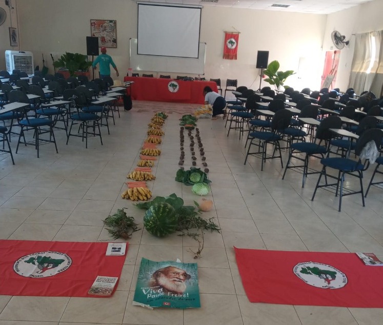 Sala de aula com bandeiras do MST espalhadas no chão e algumas frutas e legumes como melancia, banana e cenoura. Há também um cartaz de Paulo Freire